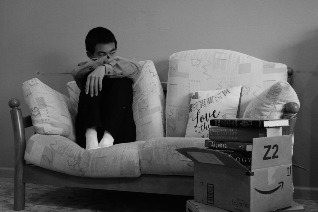 A disgruntled person sits on a loveseat with boxes of books nearby.