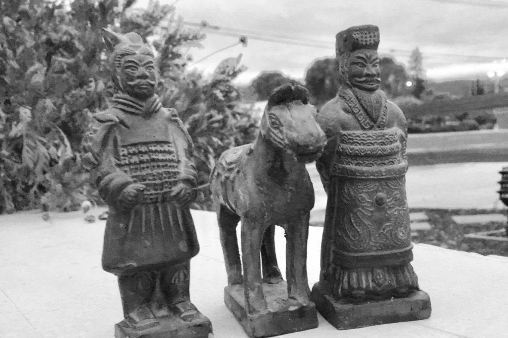 A black-and-white photo of two terracotta figures and one terracotta horse in front of foliage.